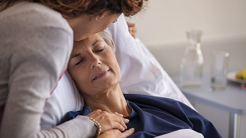 Close up of lovely daughter visiting and kissing sick mother on forehead while resting in hospital bed. Senior patient laying on gurney with eyes closed while daughter assuring a speedy recovery. Old mom patient being kissed while recovering from disease in nursing home.